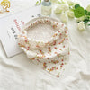 A floral patterned triangle scarf is displayed on a white cloth next to a glass bottle and a card.