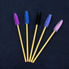 Five disposable eyelash brushes in purple, blue, black, and pink are arranged diagonally on a dark surface.