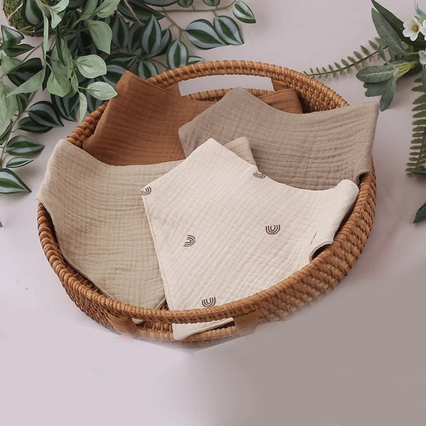 A woven basket holds a pack of three beige and brown soft cotton baby bibs with adjustable snap closures.