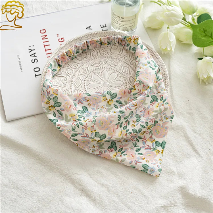 A floral patterned triangle scarf is draped next to a white card and some white roses on a light fabric.