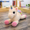 A fluffy white unicorn plush toy with pink accents and a rainbow mane sitting on a wooden floor.