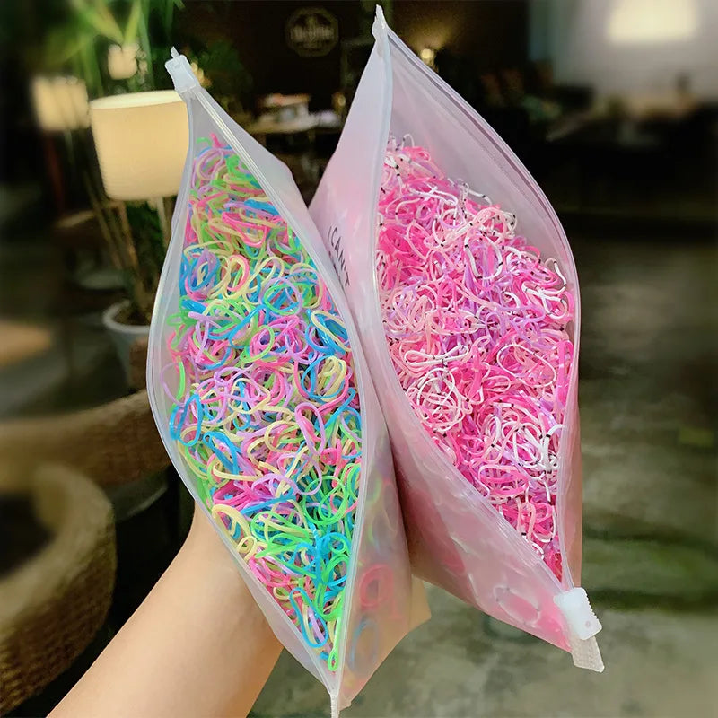 A hand holds two clear plastic bags filled with colorful disposable elastic hair bands for girls