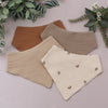 A set of three soft cotton baby bibs in brown and beige tones are displayed on a light surface.