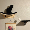 A black cat is perched on a wall-mounted cat tree with scratching stairs and hammock.