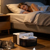 A pet water fountain device is shown on a wooden floor next to a person sleeping in bed.