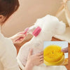 A person is using a pink silicone pet bath brush with a yellow shampoo dispenser on a white fluffy pet.