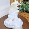 A white pet dress for a small dog featuring a bow detail and a D-ring, displayed on a wooden surface.