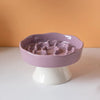 Bol en céramique violet clair à alimentation lente pour animaux, sur un piédestal blanc, fond orange.