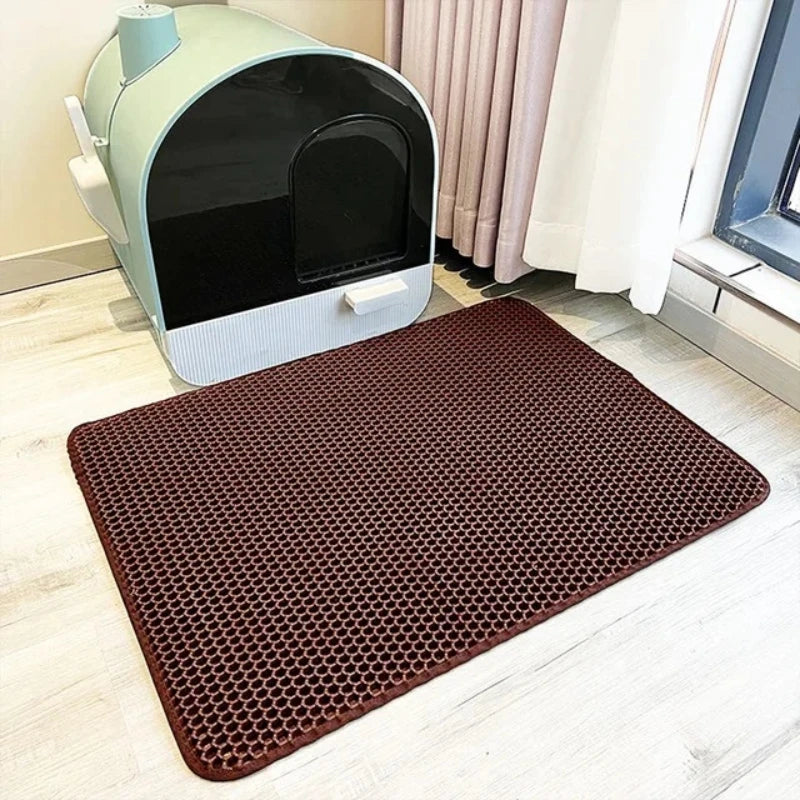 Un tapis pour litière hexagonal brun foncé est placé sur un plancher en bois clair devant une litière de chat vert clair et n