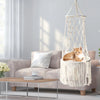 A fluffy orange cat is resting inside a bohemian macrame hammock pet bed in a modern living room.