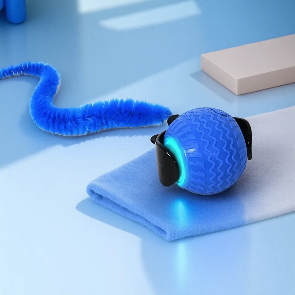 A blue automatic rotating LED pet toy resting on a white towel with a blue feather.