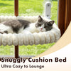 A fluffy gray and white cat is relaxing on a white cushion bed window perch with suction cups.