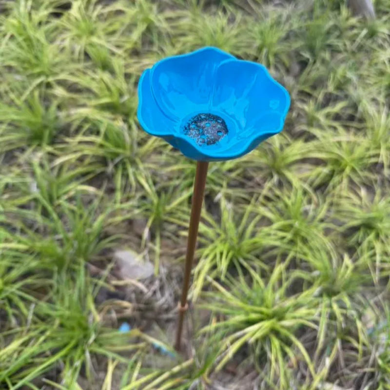A bright blue stainless steel bird feeder with a small, textured bowl is shown planted in green grass.