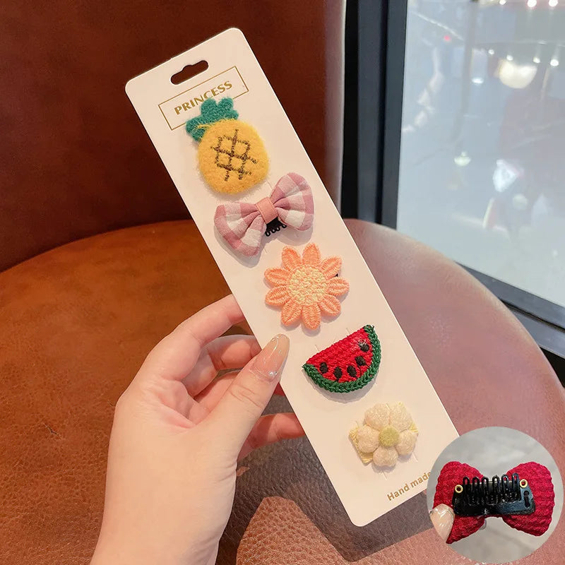 A hand holds a white card displaying five flower-shaped baby hair clips including a pineapple, bow, and watermelon slice.