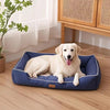 A light-colored dog is lying on a blue orthopedic dog bed indoors next to a wooden cabinet.