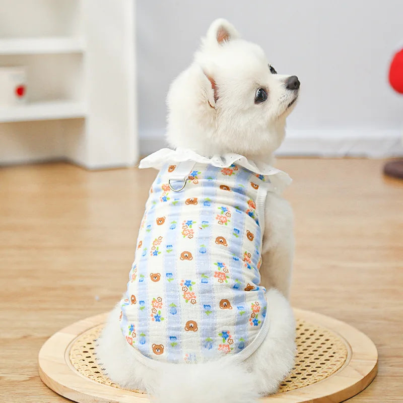 A white dog wearing a light blue cotton vest with a lace collar and cartoon print, sitting on a wooden mat.