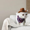 A white cat wearing a purple patterned bandana is resting on a white blanket next to an orange cowboy hat.