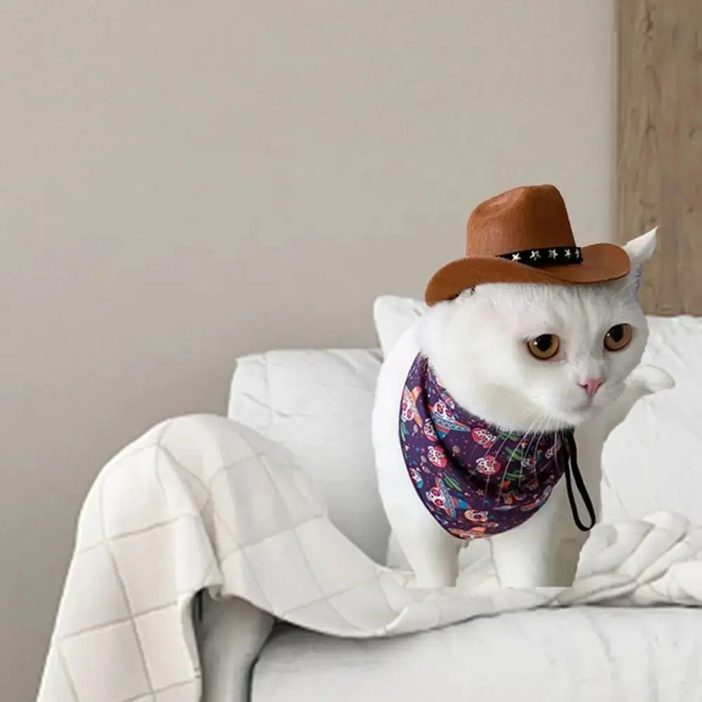 A white cat wearing a purple patterned bandana is resting on a white blanket next to an orange cowboy hat.