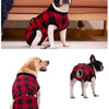 A black and white dog is wearing a red and black plaid recovery suit for post-surgery protection.