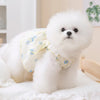 Un perro blanco esponjoso con un elegante vestido de malla floral está tumbado sobre una superficie blanca.