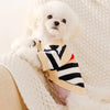 A fluffy white dog wearing a beige knit sweater with black and red stripes, resting on a cream blanket.