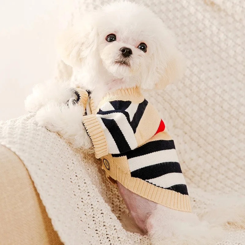 A fluffy white dog wearing a beige knit sweater with black and red stripes, resting on a cream blanket.