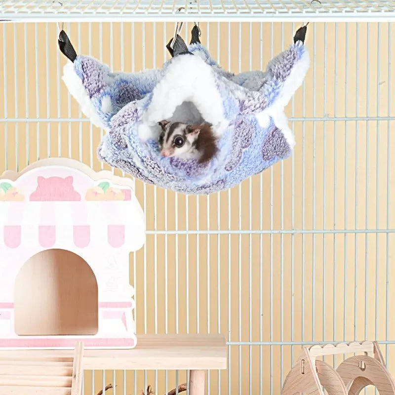 A fluffy blue and white plush pet hammock with a small kitten is hanging against a light background.