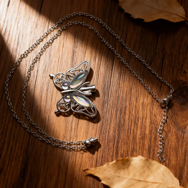 Silver butterfly ashes necklace resting on a dark wooden surface with a dried leaf.