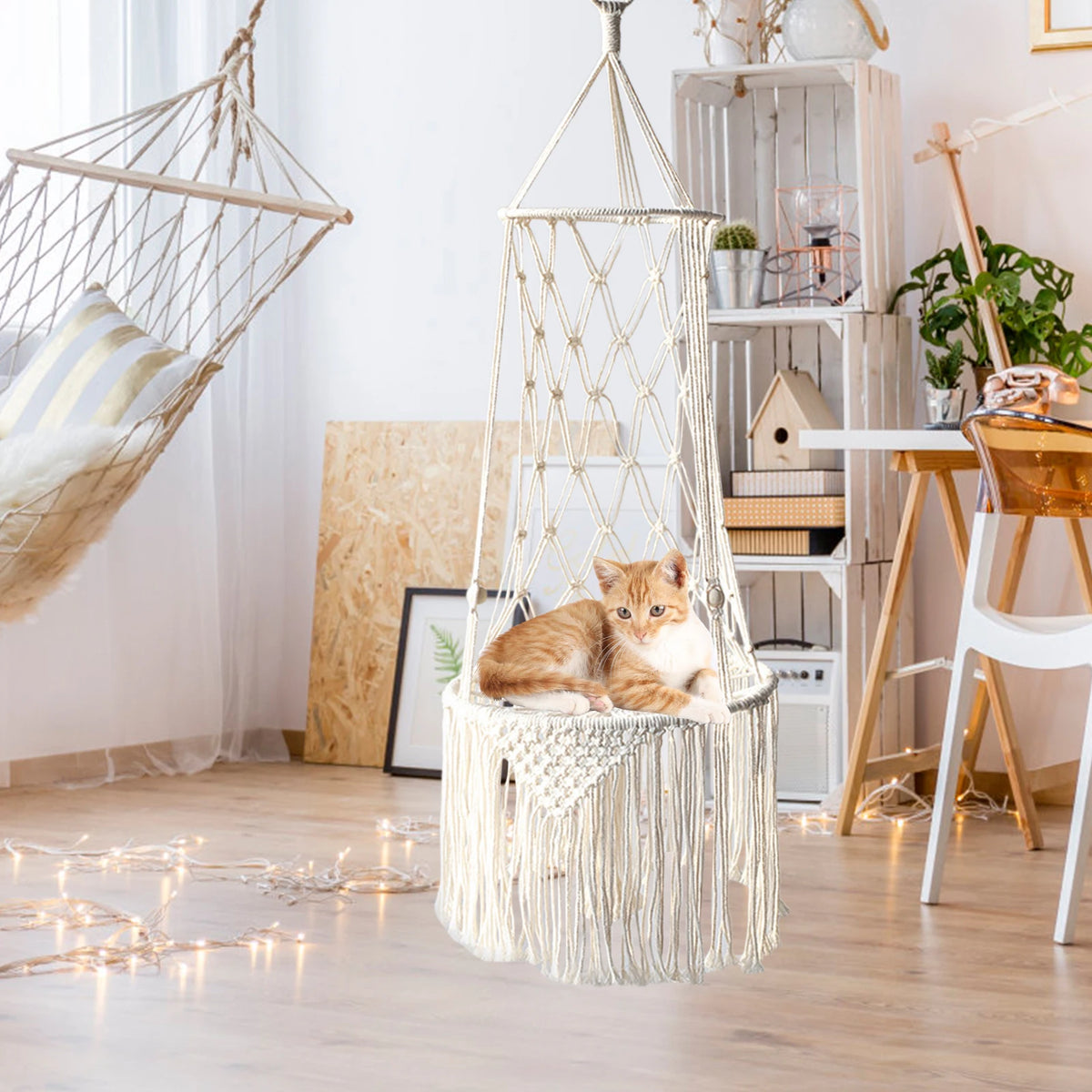 A fluffy orange cat is resting inside a large macrame hammock pet bed hanging in a bright, bohemian-style room.