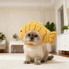 A fluffy grey cat wearing a yellow bear cap pet head cover sitting on a white rug.