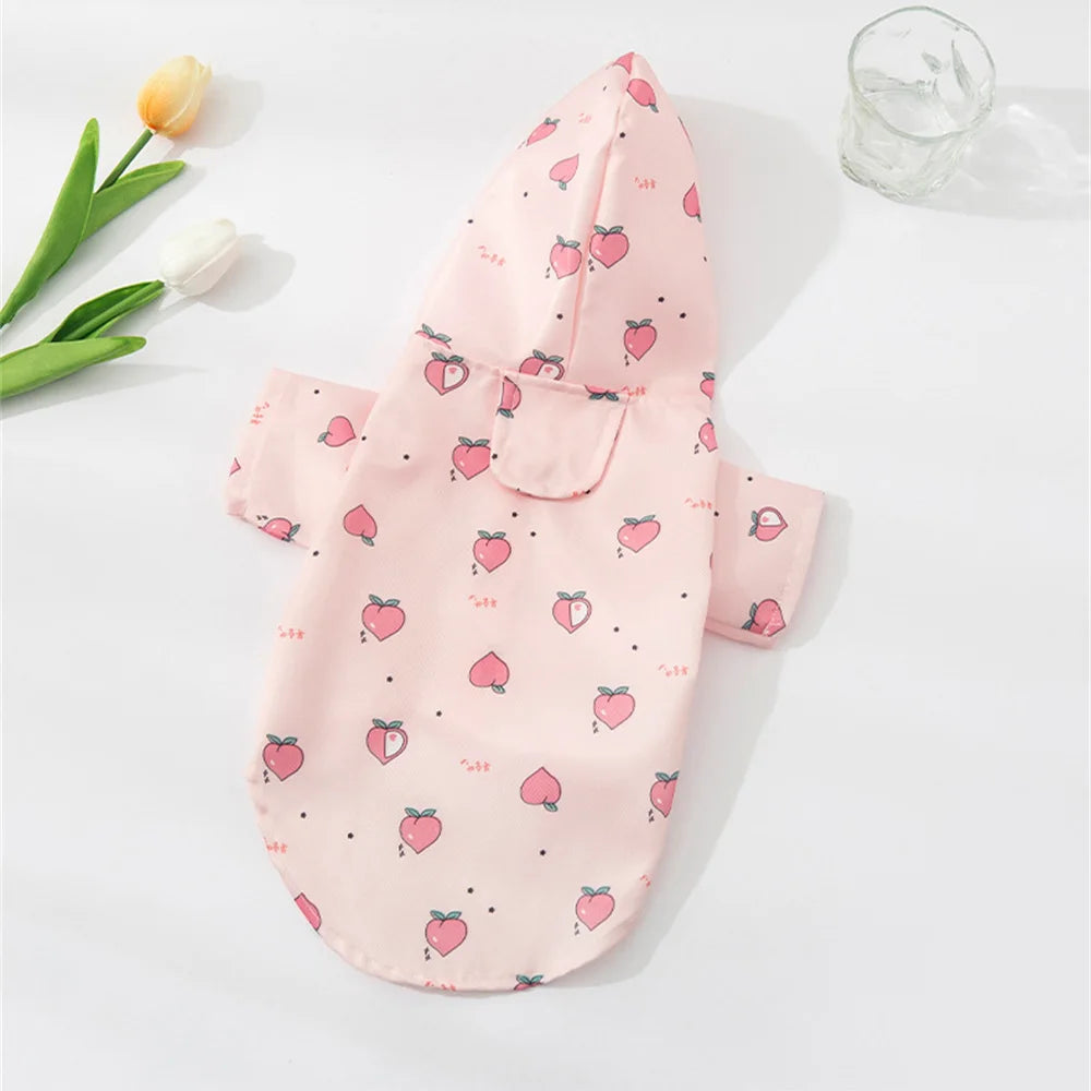 Pink cartoon printed dog raincoat for small breeds lying on a white surface with a glass and green leaves.