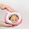 A person's hands are holding a pink plush sleeping nest with a white lining, inside of which a small brown hamster is visible