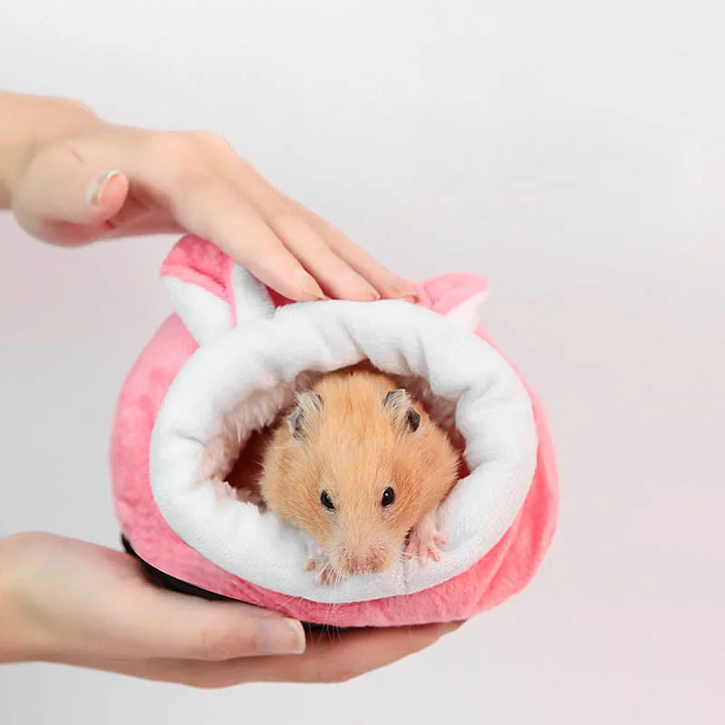 A person's hands are holding a pink plush sleeping nest with a white lining, inside of which a small brown hamster is visible