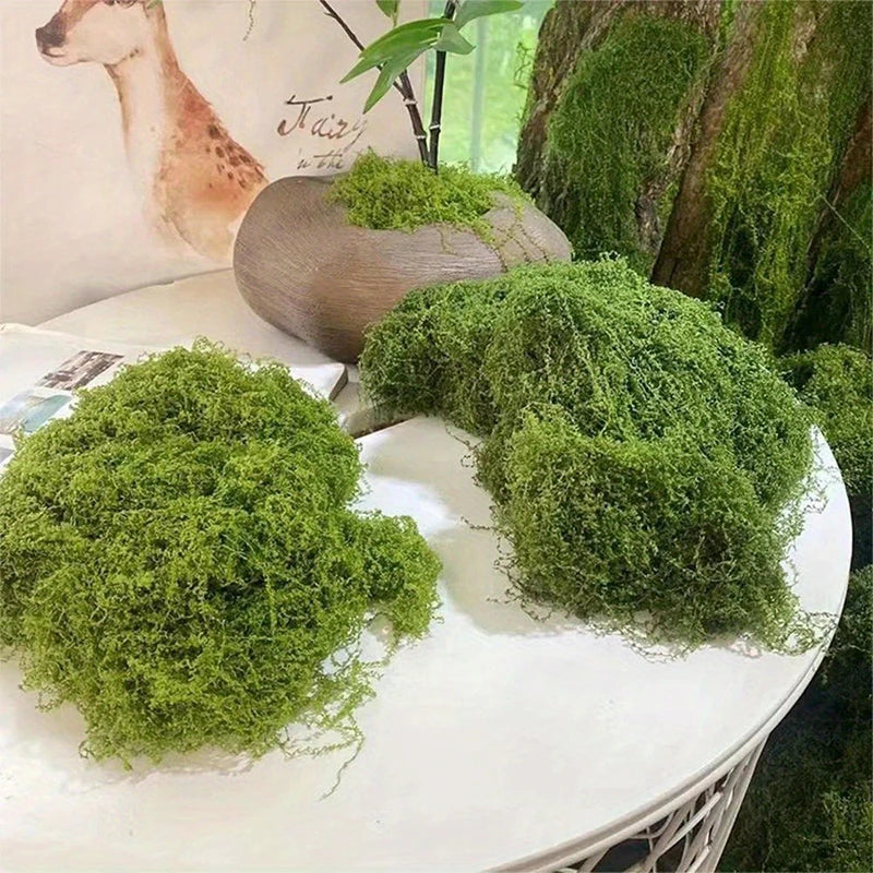 A pile of vibrant green preserved moss is displayed on a white table next to a wooden bowl and a leafy plant.
