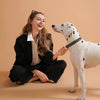 A smiling woman is gently touching the nose of a white dog wearing a black patterned collar.