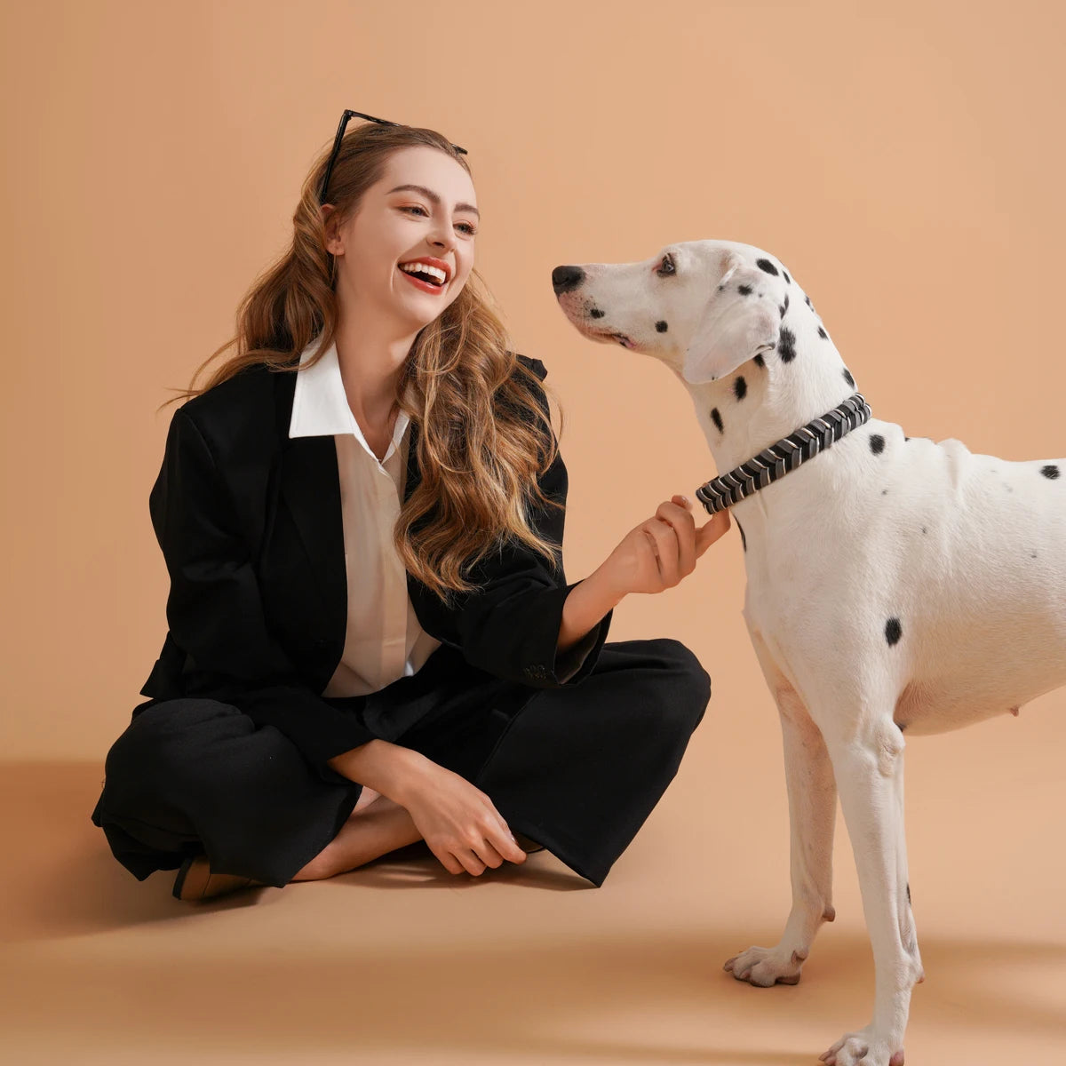 A smiling woman is gently touching the nose of a white dog wearing a black patterned collar.