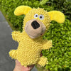 A yellow plush dog toy with floppy ears and a black nose is held against a green hedge background.