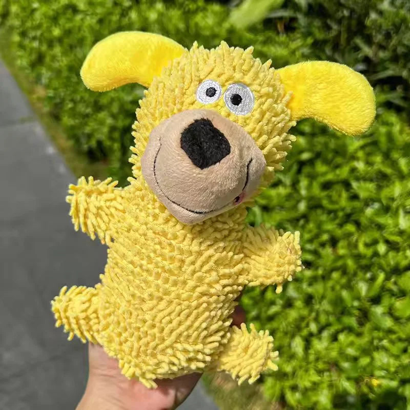 A yellow plush dog toy with floppy ears and a black nose is held against a green hedge background.