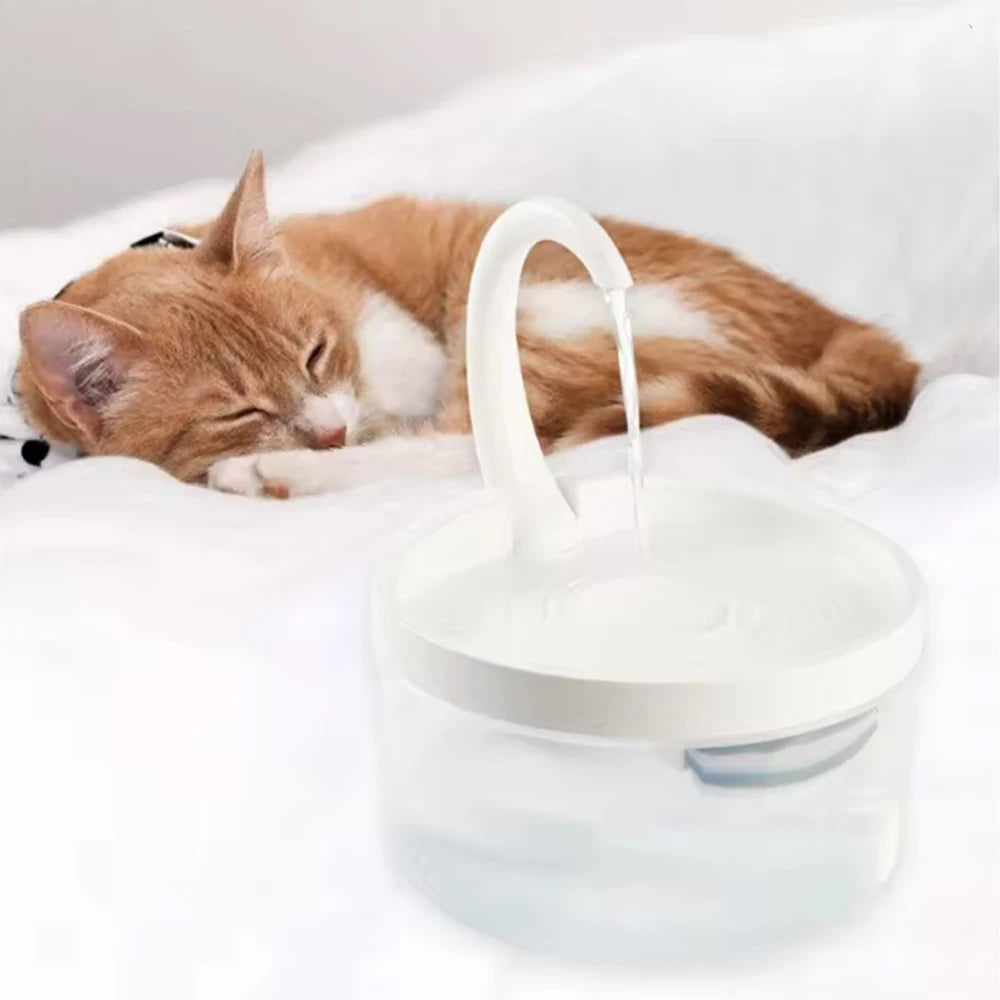 A cat is resting on a white surface near a clear water fountain with a white spout pouring water.
