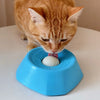 An orange cat is licking a white ball inside a blue interactive slow feeder dog bowl.