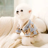 Un perro blanco esponjoso vistiendo un suéter azul claro con estampado de oso marrón, descansando sobre una manta de color cr