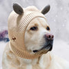 A light-colored dog wearing a beige knit ear muff with animal ears in a snowy outdoor setting.