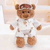 A brown teddy bear plush wearing a white outfit and an eye mask sits on a glass grid.