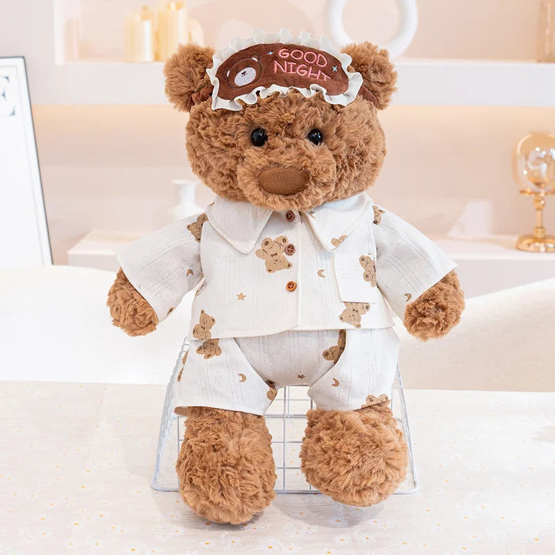 A brown teddy bear plush wearing a white outfit and an eye mask sits on a glass grid.