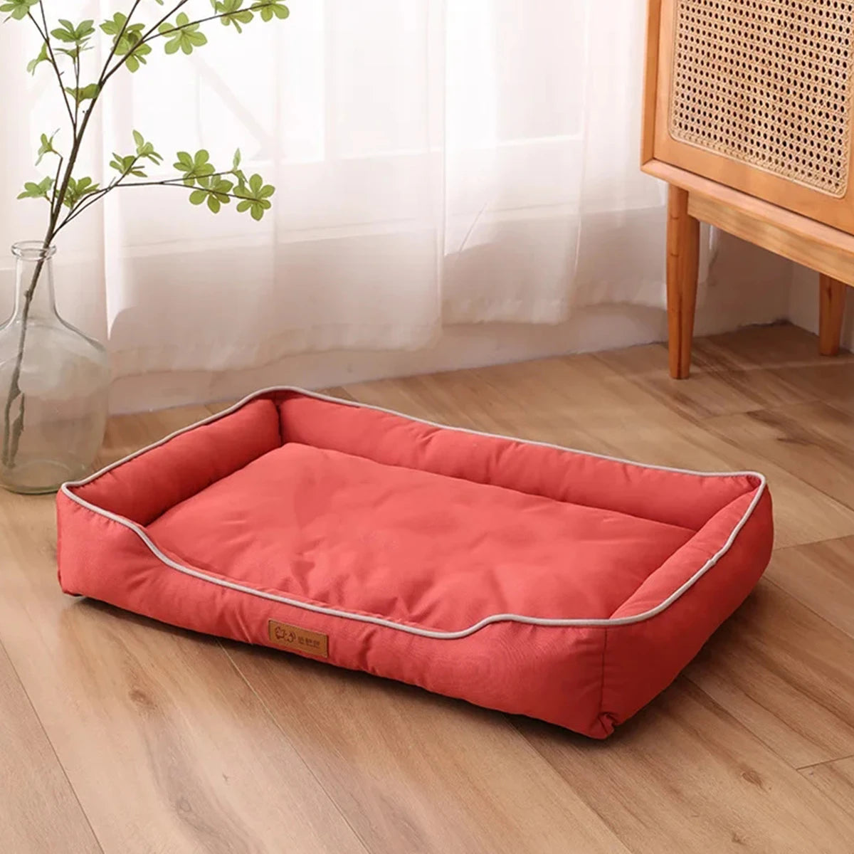 A rectangular coral orange orthopedic dog bed placed on a light wooden floor next to a window.