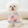 A white dog wearing a pink cartoon print vest, lying down on a light surface.
