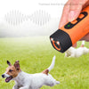 A hand holds an orange ultrasonic dog bark deterrent device, with a white dog running nearby on green grass.