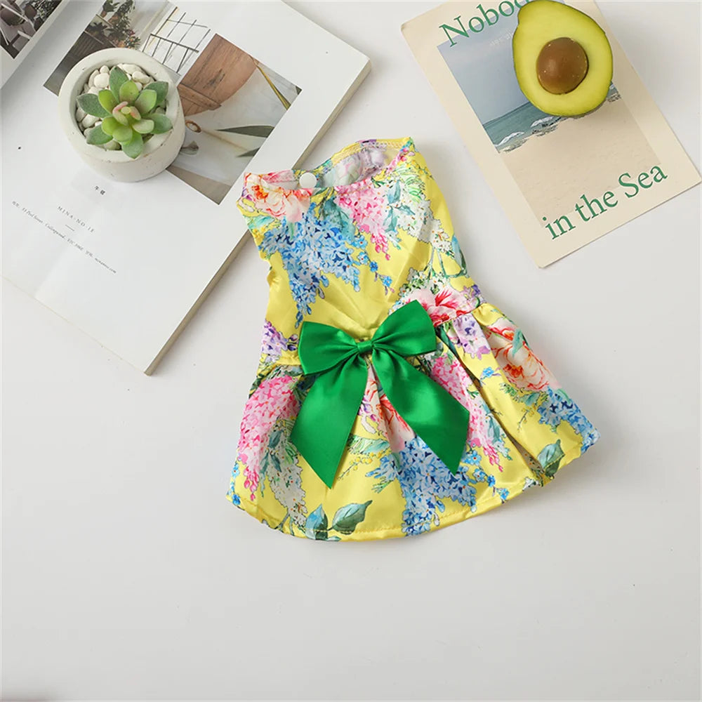 A yellow floral print dog dress with a green bow lying on a white surface.