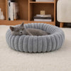 A gray cat is resting inside a round, ribbed, plush pet bed on a white carpet.