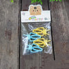 A clear plastic bag contains yellow and light blue clip-on bird cage feeders with mounting brackets on a wooden surface.